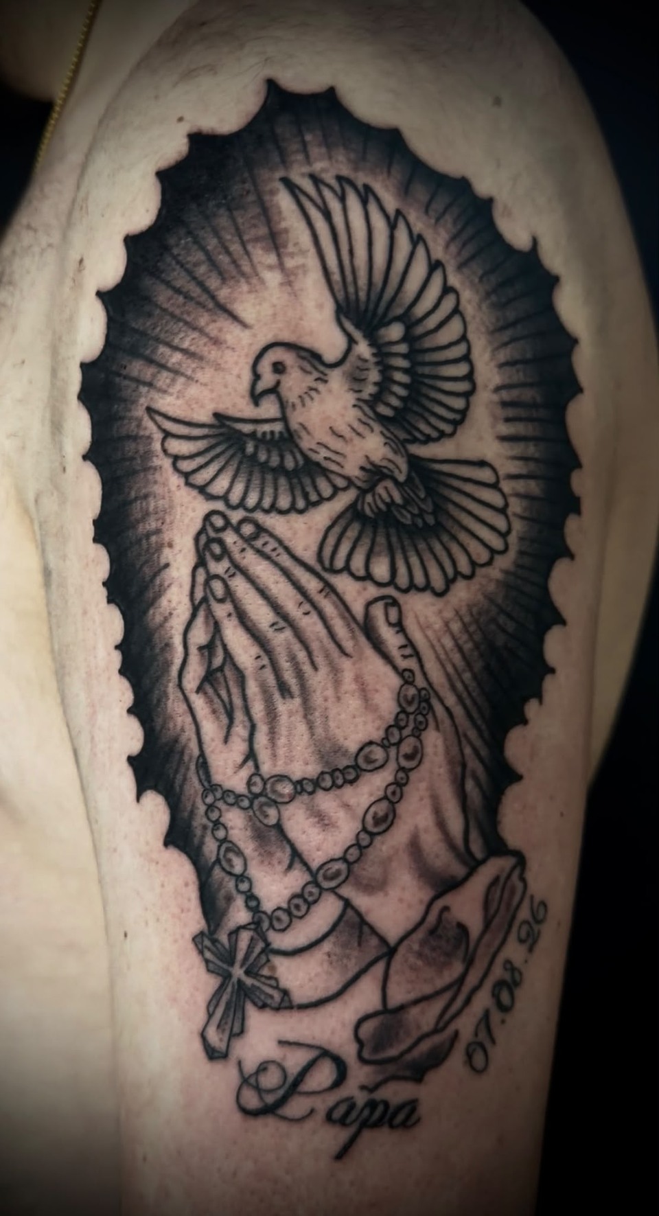 Schwarz-graues Tattoo mit einer Taube über betenden Händen, Rosenkranz und Schriftzug am Oberarm.
