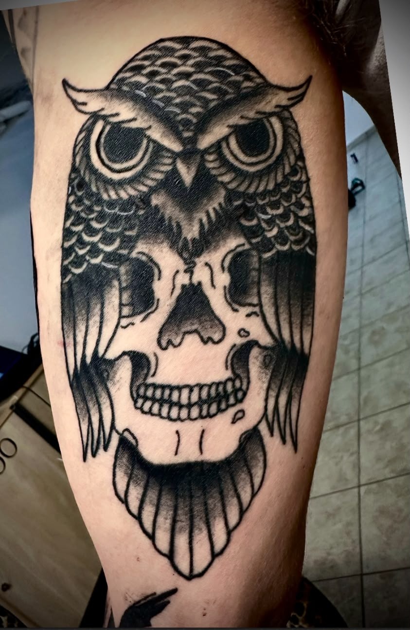 Schwarzes Tattoo einer Eule, deren Flügel einen Totenkopf umrahmen.